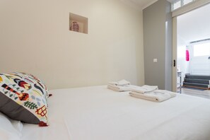 Una cama bien tendida con sábanas blancas y un cojín de colores, un pequeño adorno en la pared y vista a una escalera que conduce a una puerta.