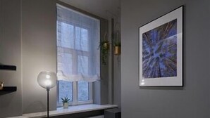 Una habitación moderna con ventana, un cuadro abstracto enmarcado, una lámpara de pie y un reposapiés verde.