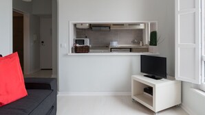 En kompakt opholdsstue med et tekøkken, et fjernsyn på et stativ og en rød pude på en sofa.