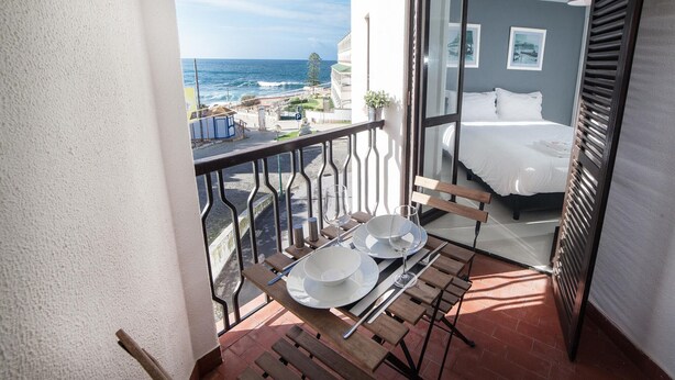Ein Balkon mit Esstisch, ein Bett mit weißer Bettwäsche und Blick aufs Meer.