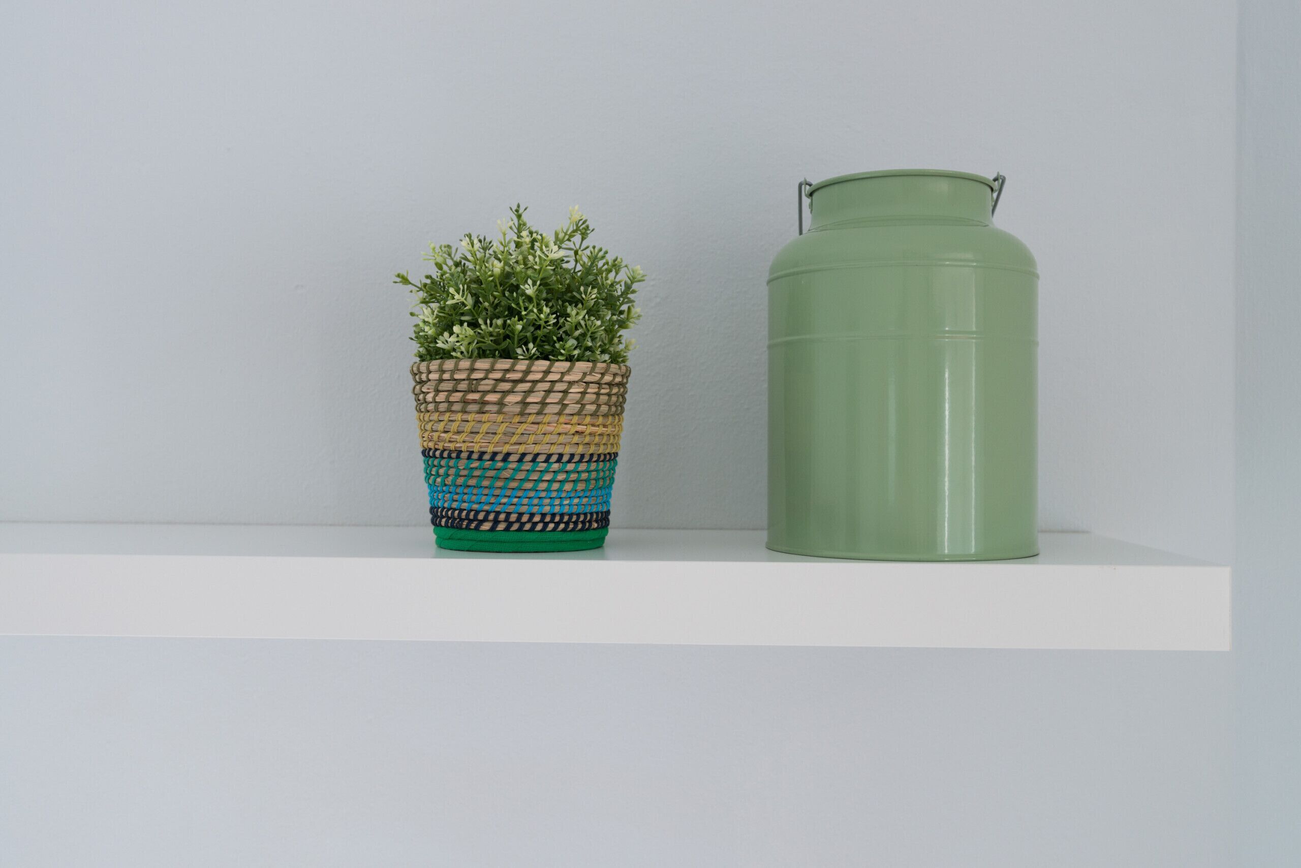 Un cesto intrecciato con una pianta e un contenitore metallico verde su uno scaffale bianco.