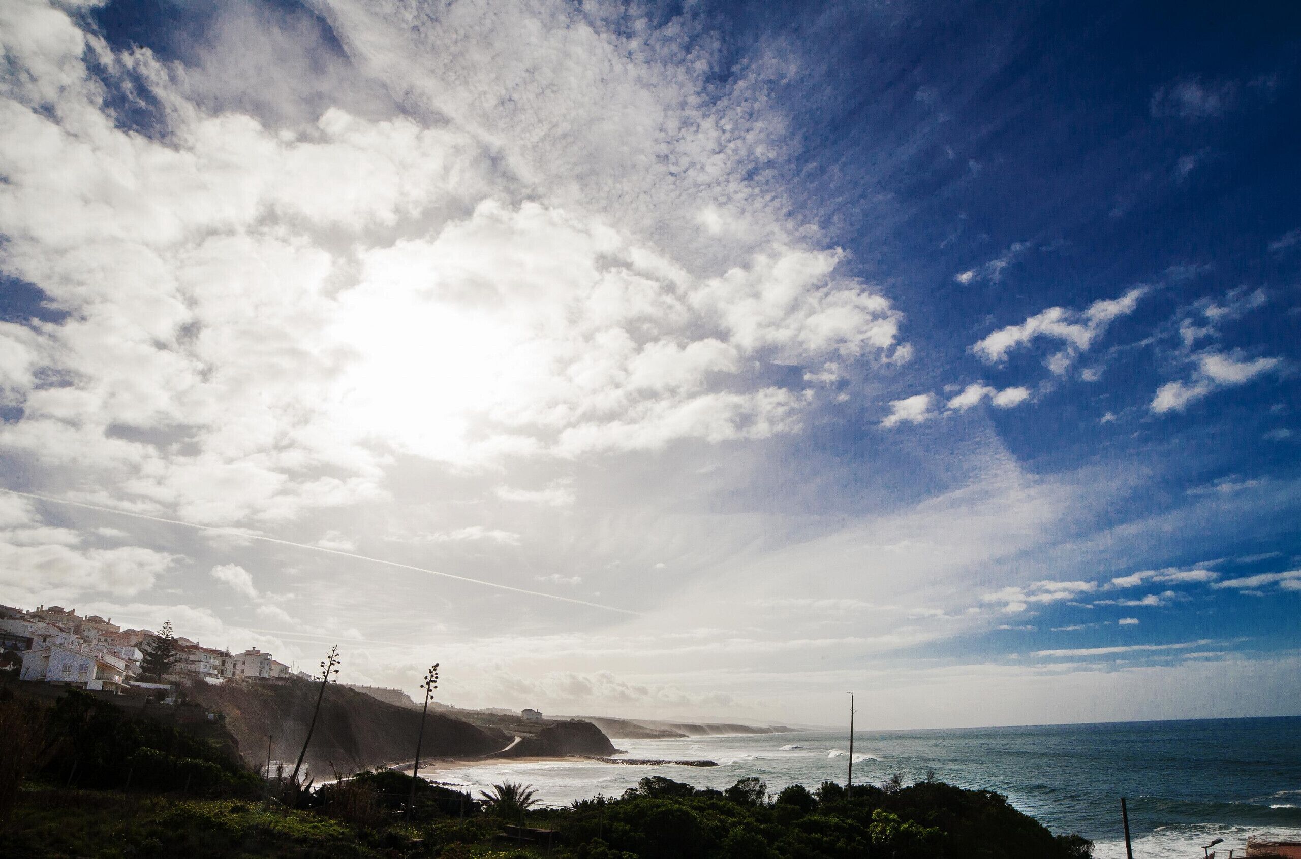 A coastal landscape with houses on a cliff, a view of the sea, and a partly cloudy sky.