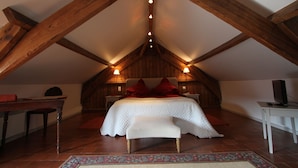 A bedroom with a sloped ceiling, wooden beams, a bed with red pillows, a bench, a table, and a television on a stand. - Le Clos Xavianne (Ribécourt-la-Tour)