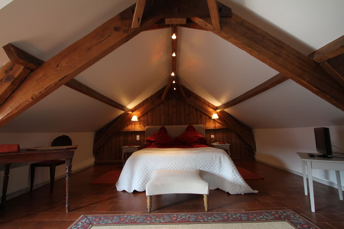 Un dormitorio con techo inclinado, vigas de madera, una cama con almohadas rojas, un banco, una mesa y un televisor sobre un soporte.