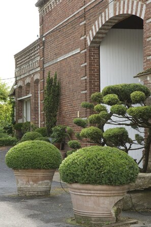 A brick building with a white door and a large arched window. There are potted topiary trees in front of the building. - Le Clos Xavianne (Ribécourt-la-Tour)