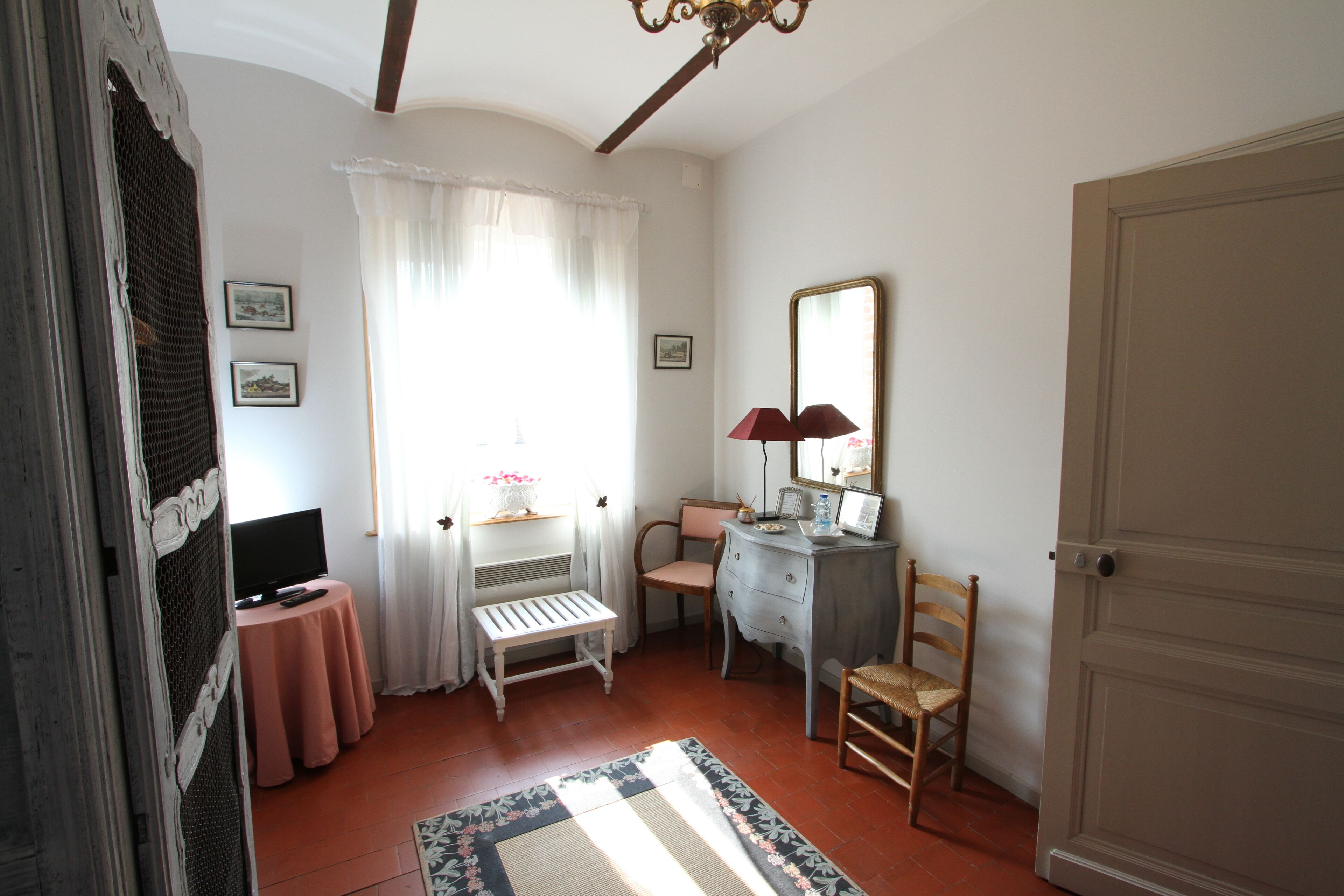 A room with a television on a small table, a desk with a lamp, a chair, a dresser, and a door.