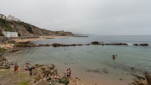 Uma praia costeira com falésias rochosas, pessoas aproveitando a água e edifícios numa encosta.