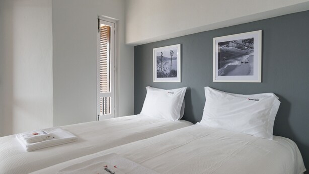 Quarto de hotel com duas camas, roupa de cama branca, uma janela com persianas e dois quadros na parede.