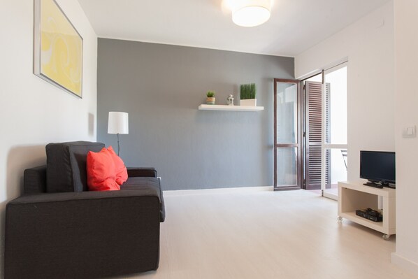 A modern living room with a grey sofa, a red pillow, a mounted TV, and a white shelf with plants.