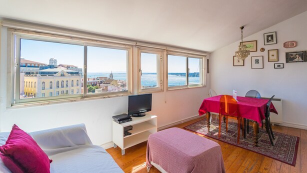 A room with a bed, a TV on a stand, a dining table with chairs, and large windows with a city view.