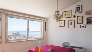 A dining area with a table set for two, a chandelier, and a view of the sea and buildings through a large window.