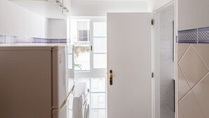 Una cocina con armarios blancos, un refrigerador y una lavadora. Hay una puerta que da a otra habitación.