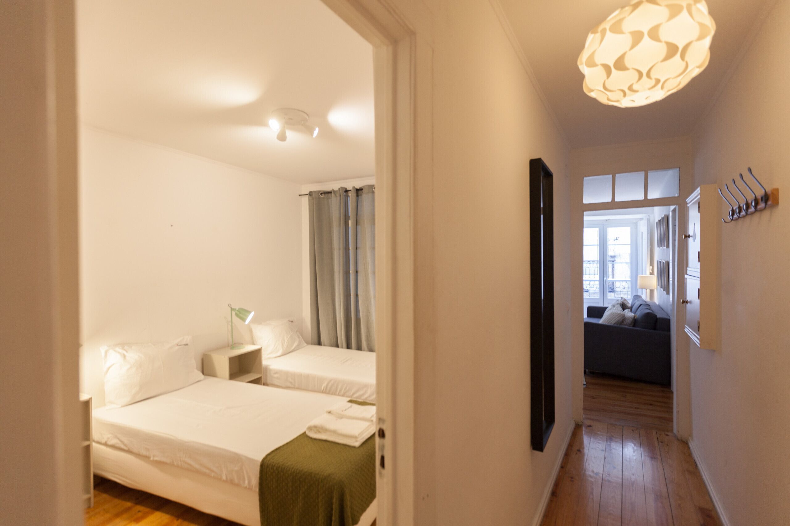 A hotel room with two beds, a hallway leading to a living area, and a modern chandelier.