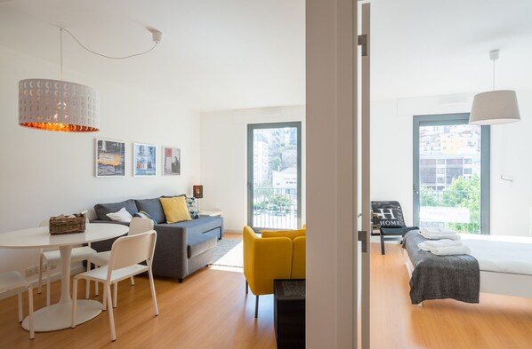 A modern living room with a sofa, a dining table, and yellow chairs. There is a bed with a grey blanket and a bench with a 'HOME' pillow.