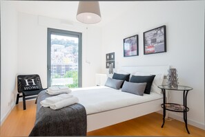 A modern hotel room with a large bed, a view of the city, and a chair with a "H HOME" sign.