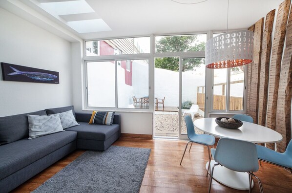 A modern living room with a grey sofa, a dining table with chairs, and a large window with a view of the outside.