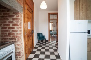 A narrow hallway with a checkered floor, a wooden door, a blue chair, and a white refrigerator.