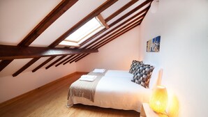 A bedroom with a sloped ceiling, wooden beams, a bed with a checkered pillow, a bedside lamp, and a painting on the wall. - FLH Lisbon Sun Terrace Graça III (Lisbon)