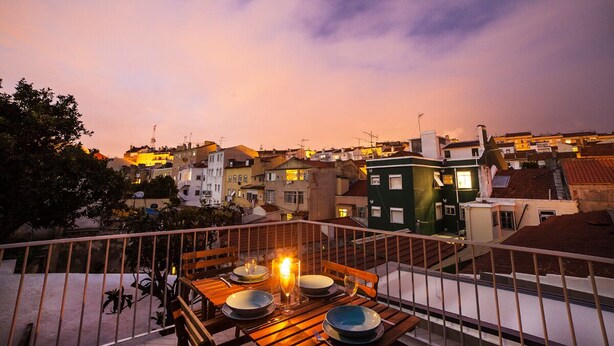 A balcony with a table set for dining, overlooking a cityscape at dusk. - FLH Lisbon Sun Terrace Graça IV (Lisbon)