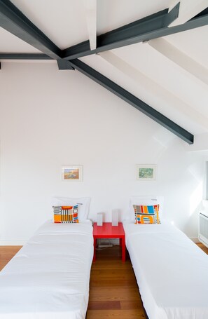 Une chambre avec deux lits simples, une table de chevet rouge, des murs blancs et un plafond en pente avec des poutres apparentes.