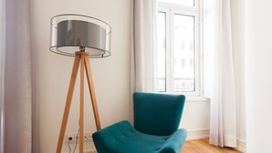 A teal armchair next to a floor lamp with a clear shade, positioned in a room with wooden flooring and a window with white curtains.