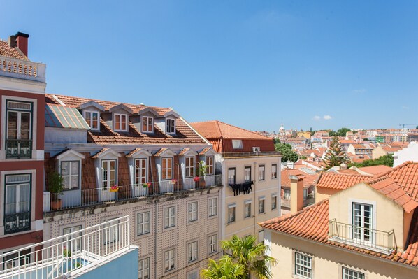 A row of traditional European buildings with red-tiled roofs, balconies with railings, and a clear blue sky.