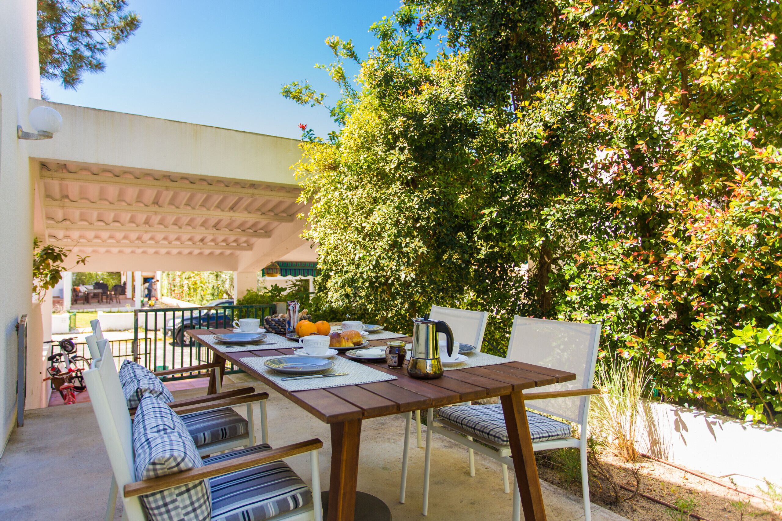 An outdoor dining area with a wooden table and chairs, surrounded by greenery and a pergola.