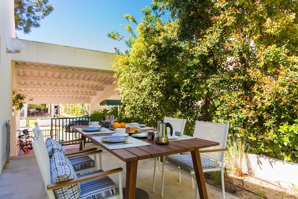 An outdoor dining area with a wooden table and chairs, surrounded by greenery and a pergola.