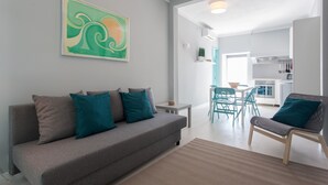 Un salon moderne avec un canapé gris, des coussins turquoise, une table basse en bois et un fauteuil turquoise. On aperçoit une cuisine à l’arrière-plan, avec des meubles blancs, ainsi qu’un coin repas avec des chaises turquoise.