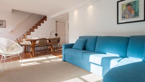 Ein modernes Wohnzimmer mit einem blauen Sofa, einem Essbereich mit Holztisch und -stühlen sowie einer Treppe, die in eine andere Ebene führt.