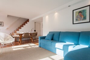 A modern living room with a blue sofa, a dining area with a wooden table and chairs, and a staircase leading to another level.