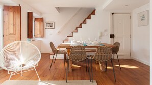 Ein Essbereich mit einem Holztisch, gedeckt für vier Personen, umgeben von Korbstühlen. Ein weißer Stuhl befindet sich links. Eine Treppe mit Holzstufen führt zu einer oberen Ebene.
