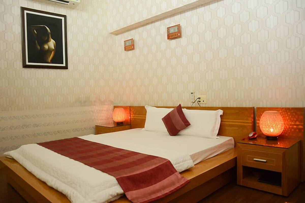 Une chambre d’hôtel avec un grand lit, deux tables de chevet, une petite table, un climatiseur fixé au mur et un tableau encadré accroché au mur.