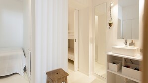Un dormitori modern amb un llit, un petit tamboret de fusta, una tauleta de nit blanca i un mirall gran sobre un lavabo blanc.