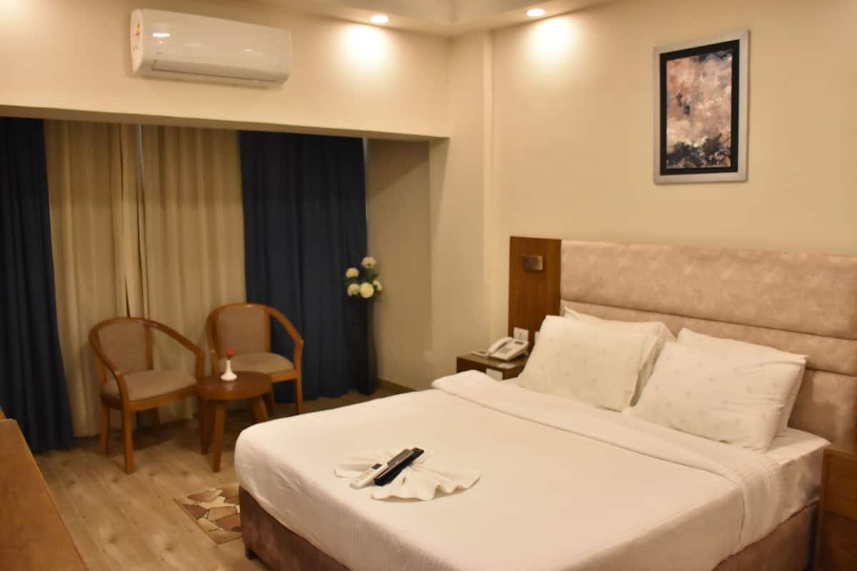 A hotel room with a bed, two chairs, a small table, an air conditioner, a nightstand, a phone, and a painting on the wall.