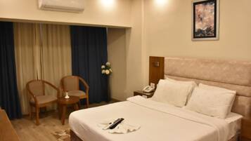 Habitación de hotel con una cama, dos sillas, una mesita, aire acondicionado, mesita de noche, teléfono y un cuadro en la pared.
