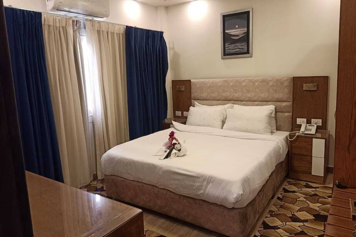 A hotel room with a bed, a bedside table, a nightstand with a phone, a window with curtains, and a wall-mounted air conditioner.