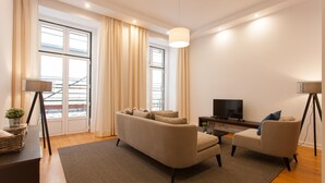 Una sala de estar moderna con un sofá, una mesa de centro y un televisor. Hay un ventanal con cortinas, una lámpara y una planta pequeña sobre una mesita.