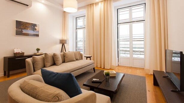 Una sala de estar moderna con sofá, una mesa de centro, televisor y ventanas amplias con cortinas.