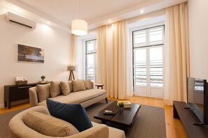 Ein modernes Wohnzimmer mit Sofa, Couchtisch, Fernseher und großen Fenstern mit Vorhängen.