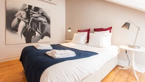 Uma cama bem arrumada com almofadas brancas e vermelhas, um cobertor azul e uma toalha dobrada. Há uma luminária moderna sobre uma pequena mesa branca ao lado da cama. Um quadro abstrato grande está na parede acima da cama.