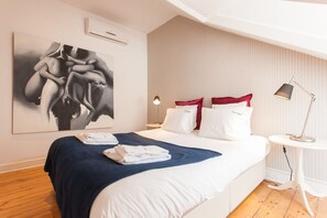 A neatly made bed with white and red pillows, a blue blanket, and a folded towel. There is a modern lamp on a small white table beside the bed. A large abstract painting is on the wall above the bed.
