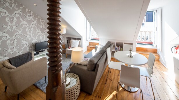 Um apartamento loft moderno com pé-direito alto, área de jantar, sala de estar com sofá e poltronas, e uma televisão ao fundo.