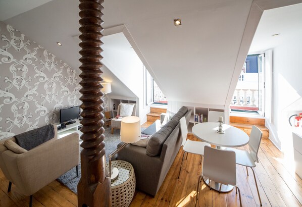 A modern loft apartment with a high ceiling, a dining area, a living room with a sofa and chairs, and a television in the background.