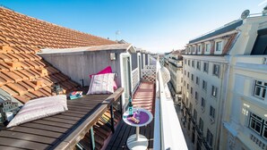 Terraço no telhado com deck de madeira, uma mesa redonda pequena com uma garrafa e copos, e uma almofada rosa.
