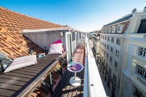 A rooftop terrace with a wooden deck, a small round table with a bottle and glasses, and a pink pillow.