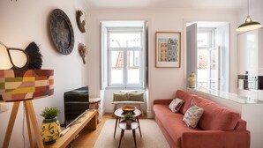 A living room with a sofa, a television, a small table, and a window with a view of buildings. - FLH Baixa Modern Place (Lisbon)