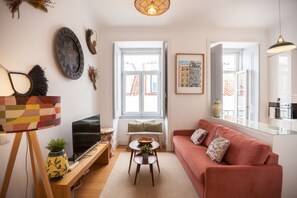 Un salon comprenant un canapé, un téléviseur, une petite table et une fenêtre donnant sur des immeubles.