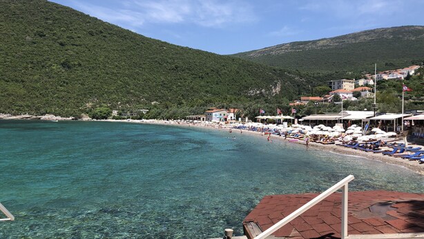 A coastal area with clear turquoise water, a beach with umbrellas and sun loungers, and buildings in the background. - Apartment for 2 guests with sea view in Herceg Novi (Meljine)