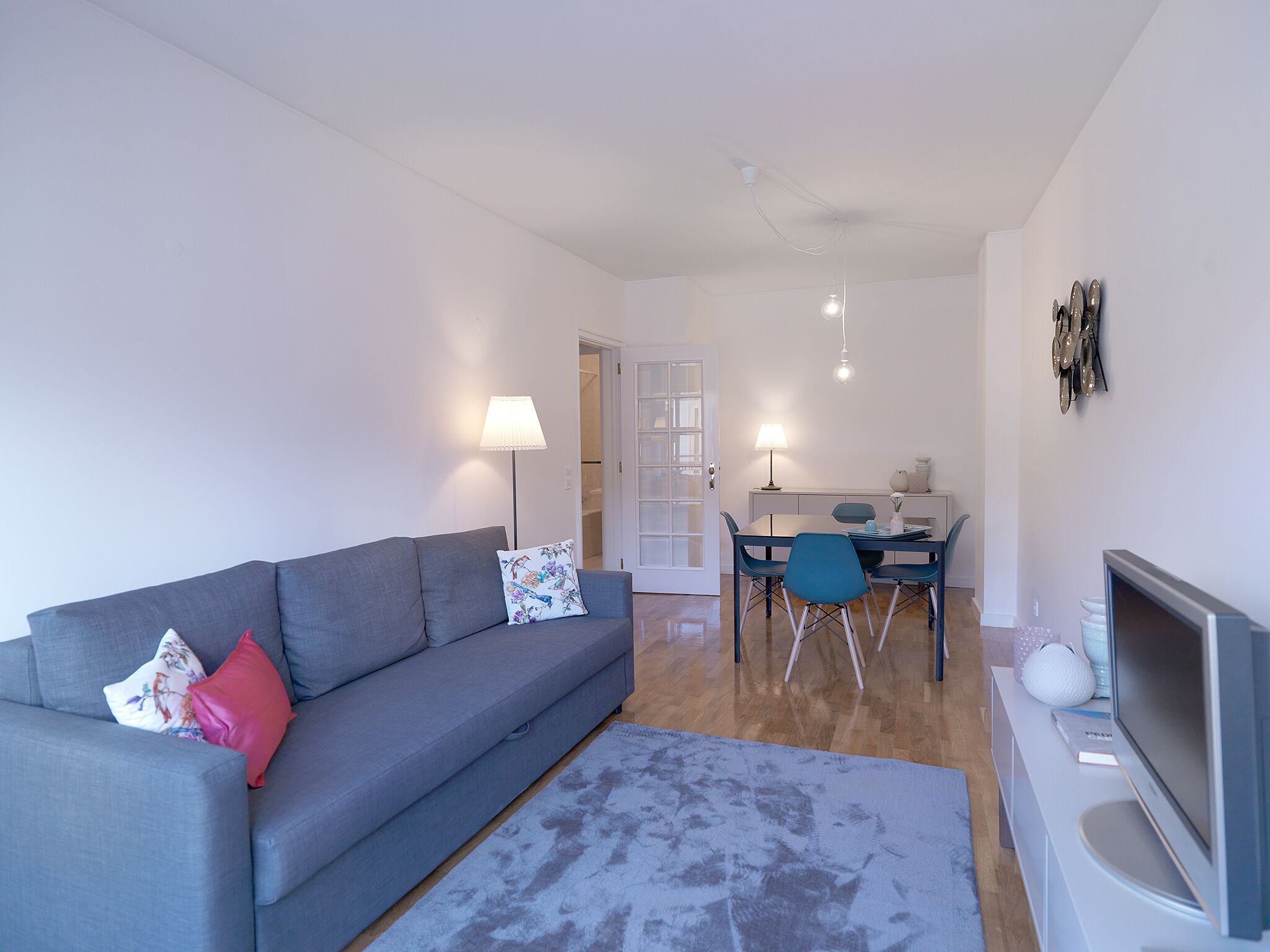 Ett modernt vardagsrum med en grå soffa, ett matbord med stolar, en tv och en lampa.